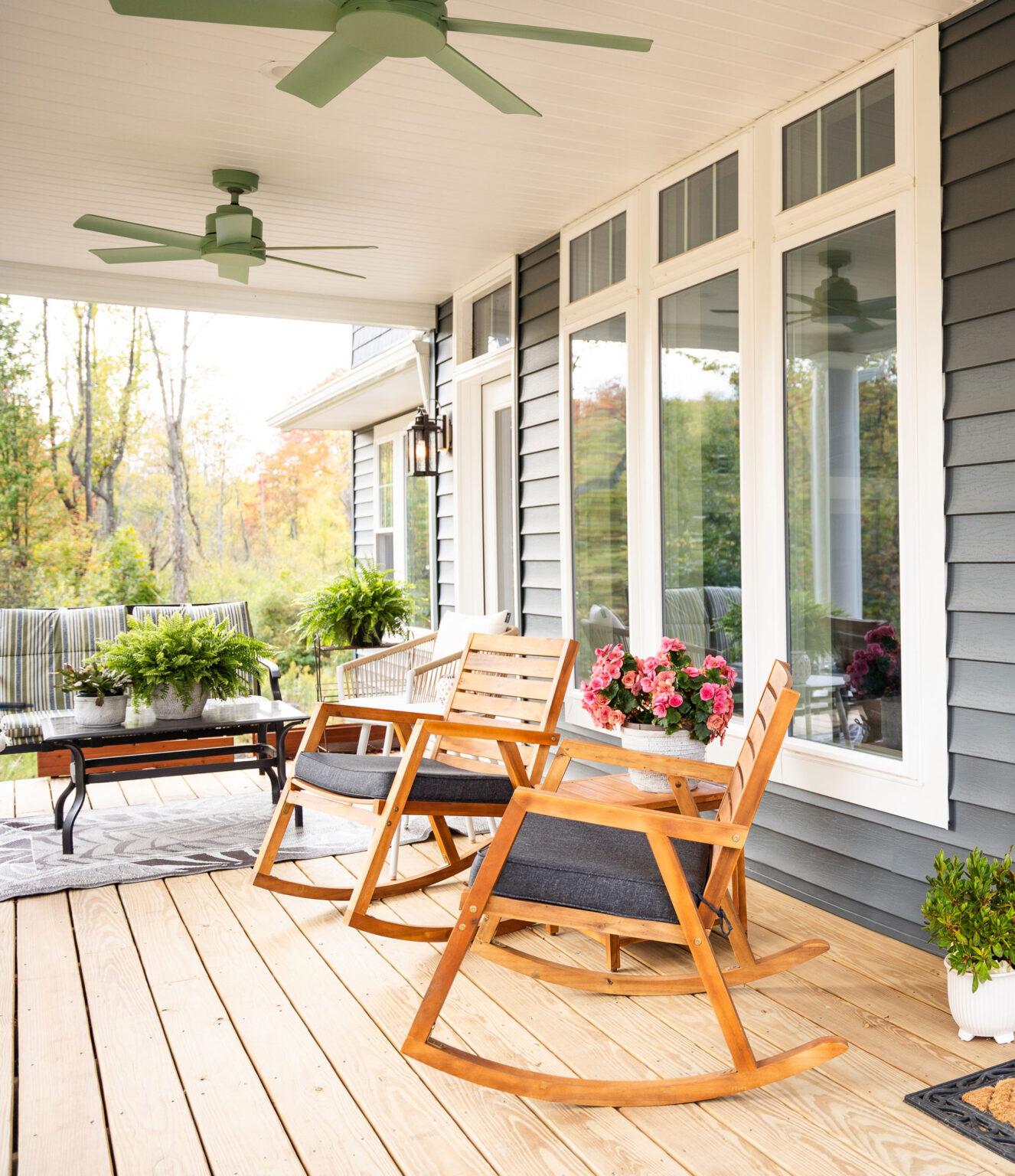 A cozy porch with two wooden rocking chairs and a small table topped with pink flowers. The area features large window casements typical of St. Louis, complemented by ceiling fans, a sofa, potted plants, and natural outdoor scenery in the background.