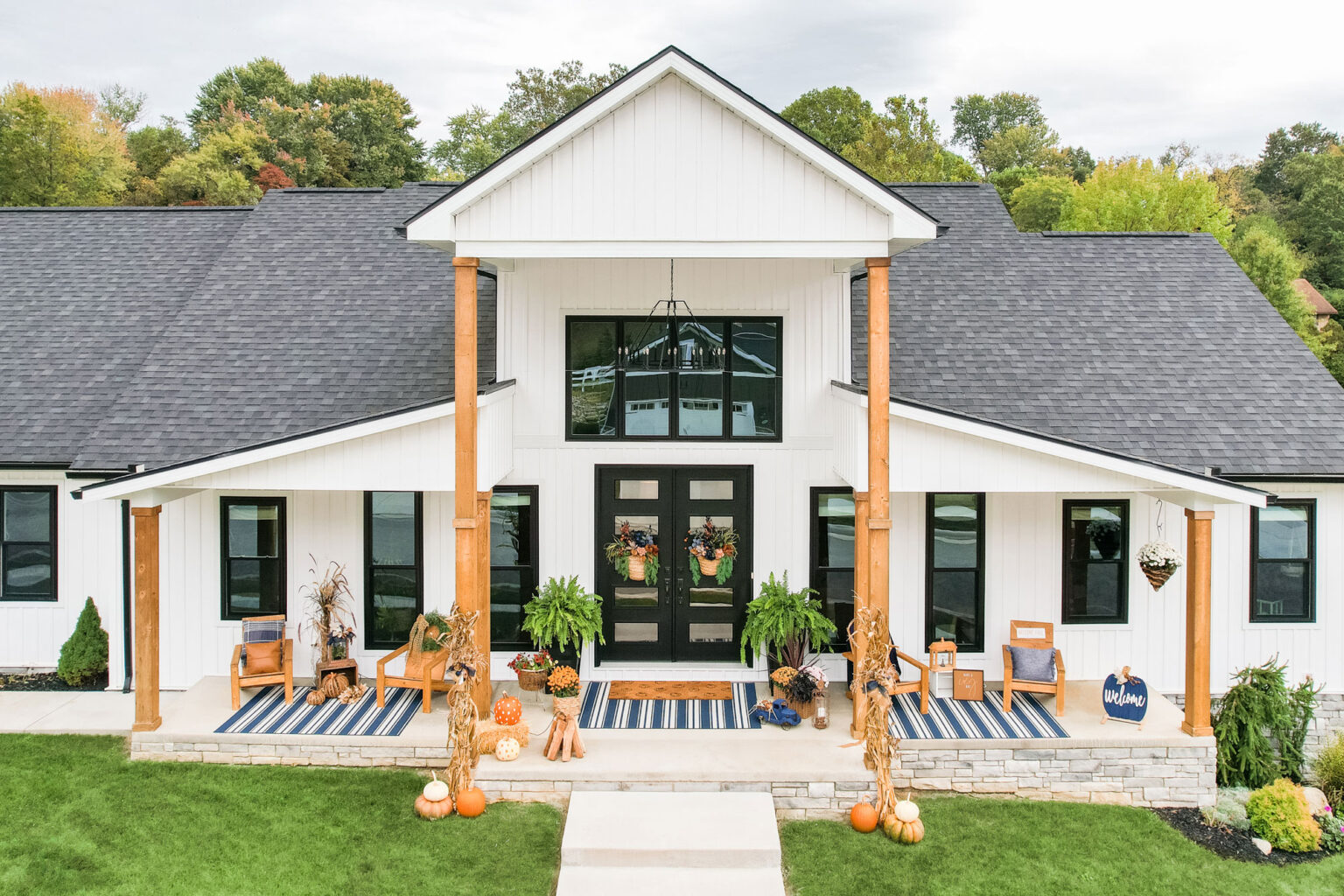 A modern white house with a black roof is decorated for autumn. The entrance features double hung windows, two black doors, pumpkins, cornstalks, and wreaths. Two rocking chairs sit on the porch, while trees with fall foliage surround this St. Louis home.