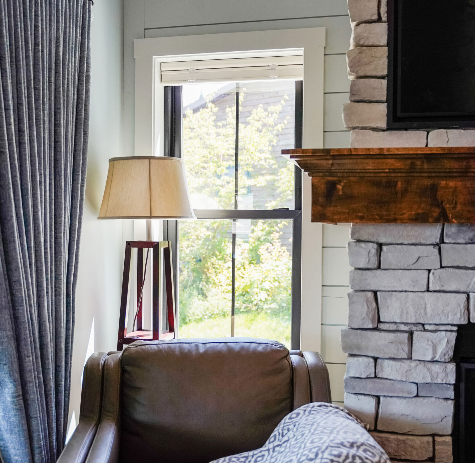 A cozy room with a leather armchair next to a stone fireplace. A tall lamp with a beige shade stands by the double-hung windows, offering a view of green trees outside. A blue curtain partially covers the window on the left, providing just the right ambiance for relaxation.