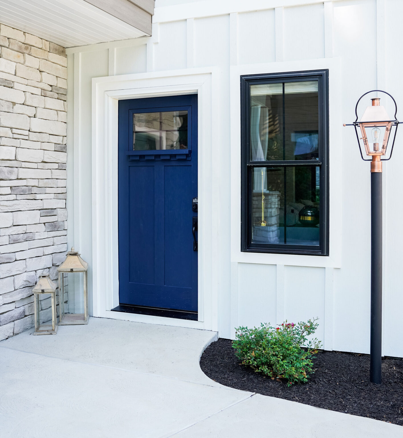 A modern entryway featuring a deep blue front door with a black-framed window beside it, reminiscent of double hung windows. The white exterior wall boasts stone accents. Two lanterns are on the ground, and a tall lamp post stands nearby, adding charm akin to classic St. Louis style.