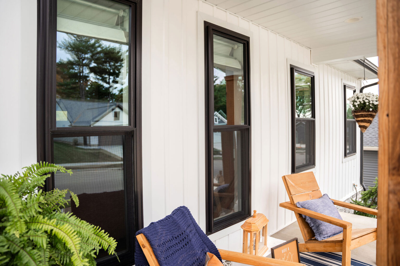 A cozy porch with two wooden chairs, each adorned with a blue cushion, and a view enhanced by the nearby double hung windows. A small wooden table sits between the chairs, and a hanging plant basket is on the right. The porch overlooks a suburban St. Louis neighborhood with trees and houses.