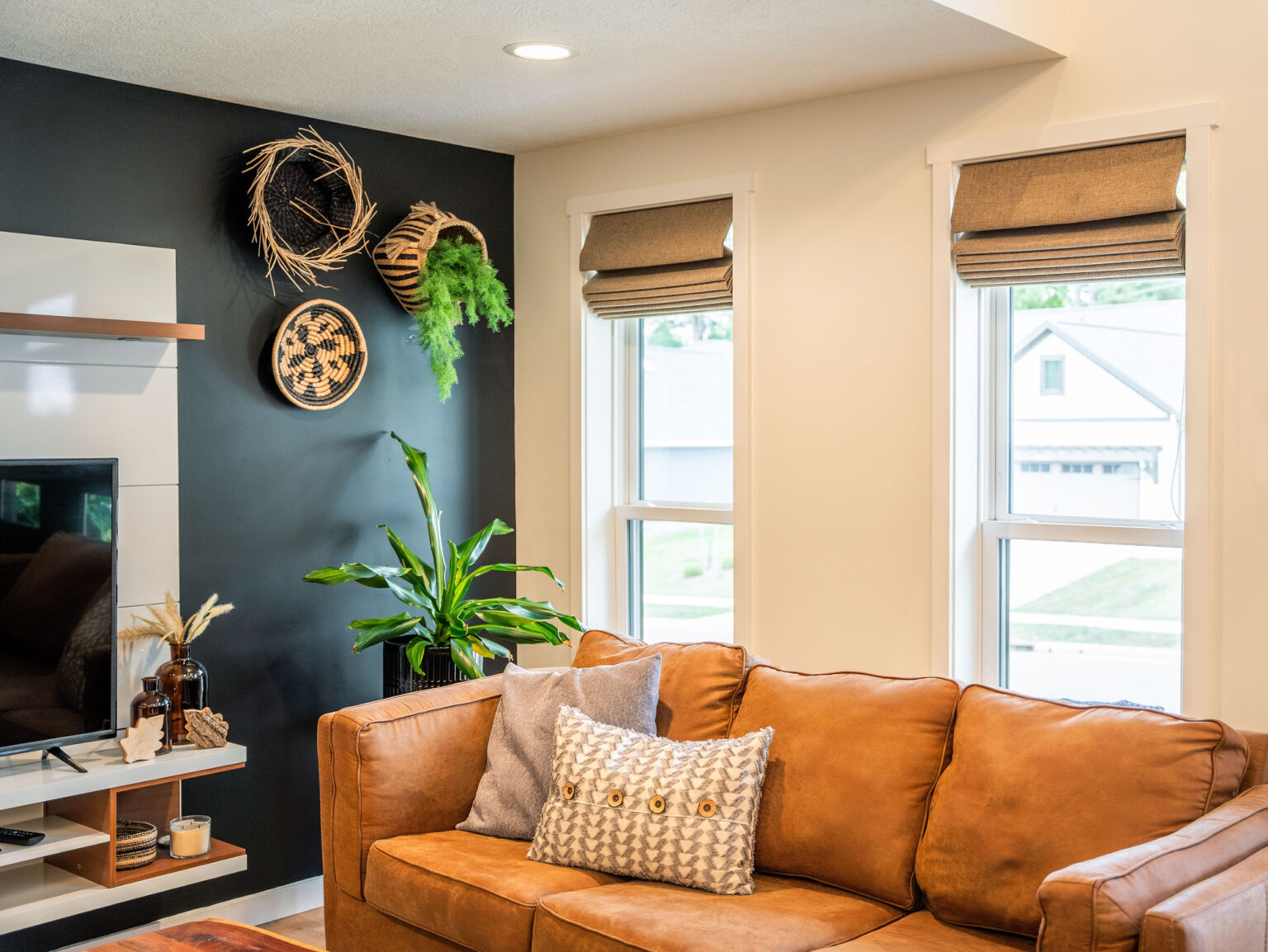 A cozy living room featuring a brown leather sofa with patterned pillows, a dark accent wall adorned with woven baskets and a plant, large double hung windows with brown roman shades framing the St. Louis skyline, and a mounted TV on a wooden shelf decorated with cherished items.