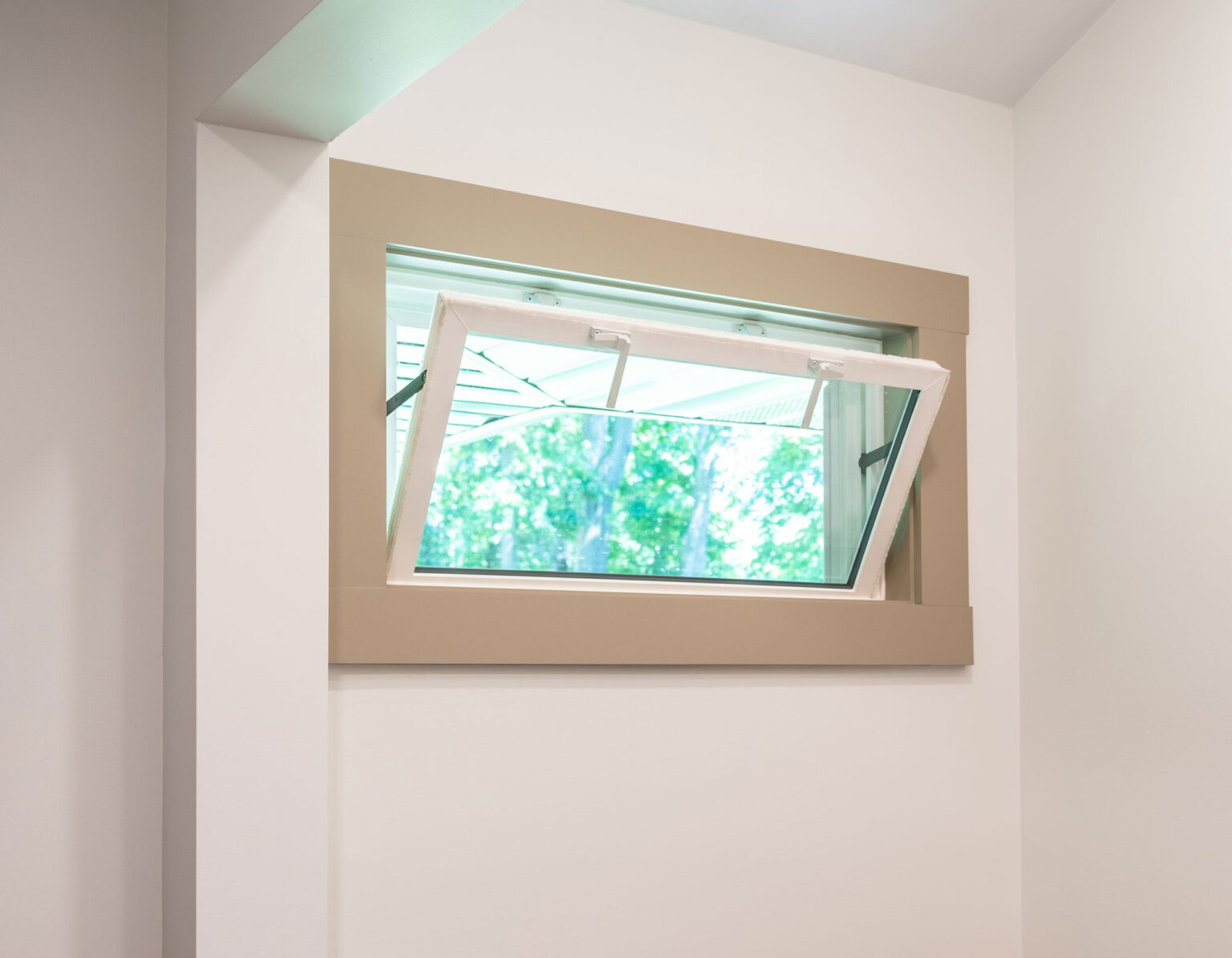 A small rectangular window with a tan frame opens outward, revealing a view of green trees and blue sky outside. Reminiscent of traditional hopper windows, this design is popular in St. Louis homes, set in a white wall to provide ample natural light to the interior space.