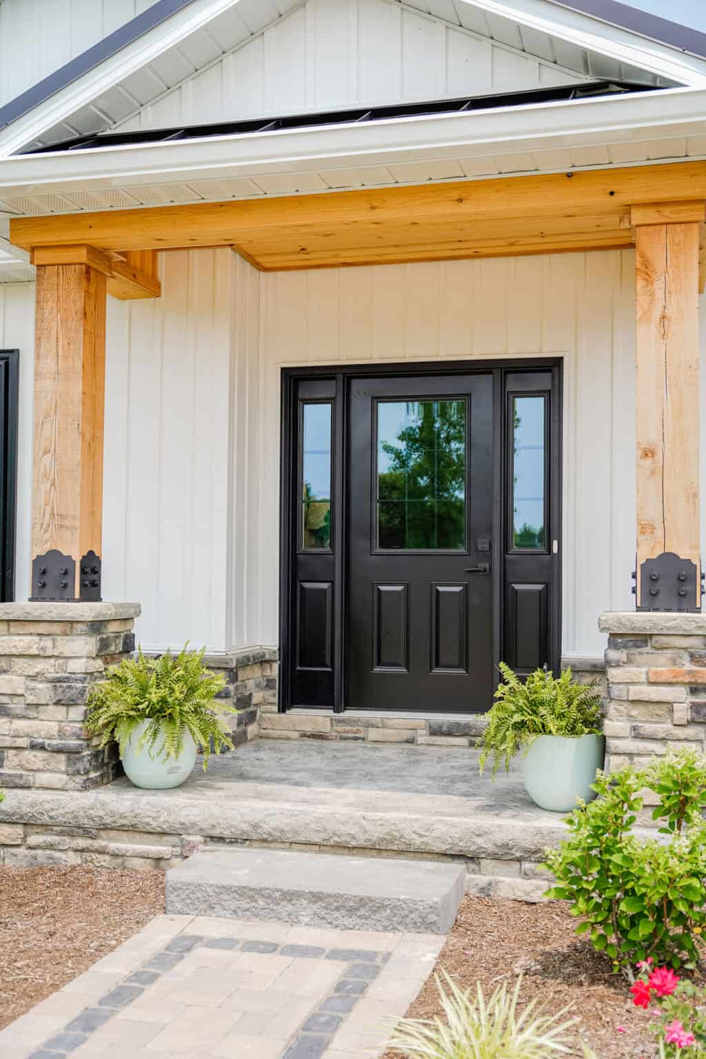 A modern home's entrance featuring a black front door with glass panels, reminiscent of the elegant entry doors seen throughout St. Louis. The doorway is framed by wooden beams and stone pillars. Two green potted plants sit on either side of the steps, with a few shrubs visible in the foreground.