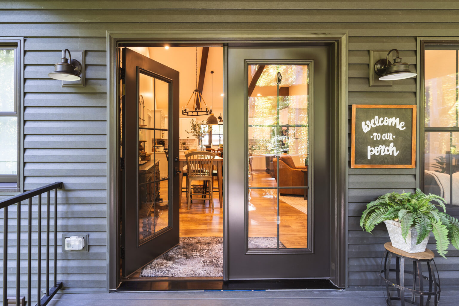 A welcoming porch entrance with open black double doors from Entry Doors St. Louis leads to a cozy interior. A sign reads "Welcome to our porch" next to a potted fern. Inside, wooden furniture and a rug create a warm atmosphere. Two wall sconces flank the doorway.