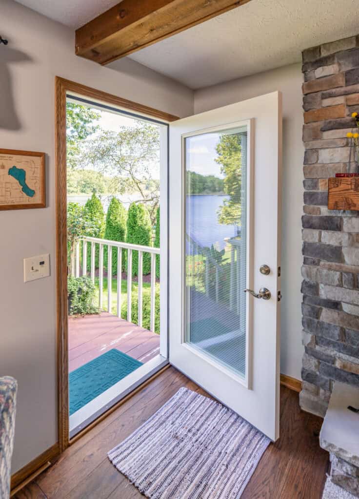 A white entry door with a glass panel opens to a view of a deck overlooking a tranquil lake surrounded by trees. The room has a stone fireplace, wooden beams, a small rug, and wall decor, creating a cozy interior that echoes the charm of Entry Doors St. Louis craftsmanship.