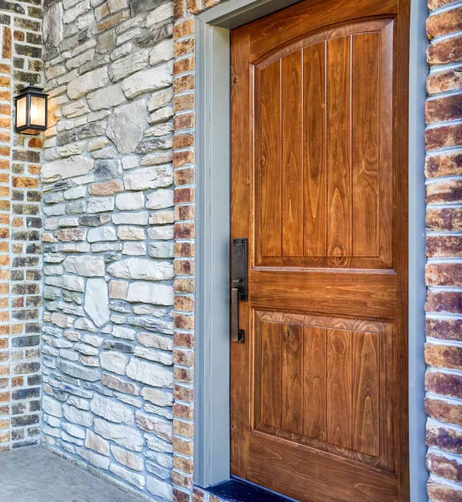 Wooden front door with a carved design set in a brick and stone wall. A black handle and lock are on the right. A black lantern-style light fixture is mounted on the left side of the doorway, epitomizing the charm of exterior doors St. Louis residents prefer.