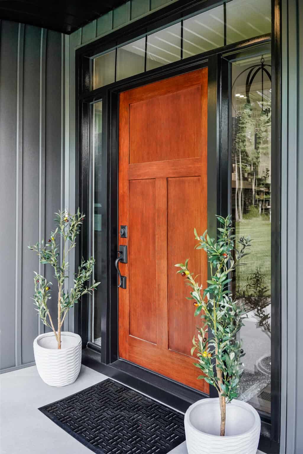 An inviting front entrance features a wooden door with glass panels, framed by dark trim. Two potted plants in white planters are symmetrically placed on either side. A black doormat lies in front on the concrete porch, showcasing elegant exterior doors St. Louis style.