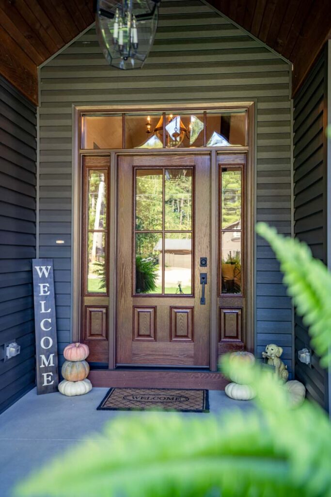 A wooden front door with glass panels is surrounded by dark siding, a testament to the elegance of exterior doors St. Louis offers. A welcome sign and stacked pumpkins grace the left, while a welcoming mat completes the porch. Ferns frame this picturesque scene with a chandelier visible through the upper window.