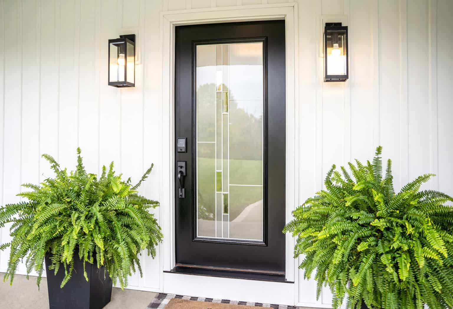 A modern black front door with a vertical glass panel featuring geometric design is flanked by two potted ferns. The white wall, adorned with rectangular black lanterns, illuminates this chic entryway often found in stylish homes across St. Louis.