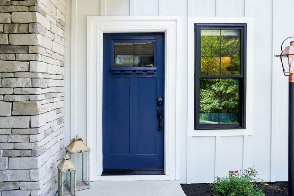 A navy blue front door with a small window at the top is set in a white paneled exterior wall. A black-framed window complements the Entry Doors St. Louis design on the right, while modern lanterns and a small shrub add charm near the stone wall on the left.