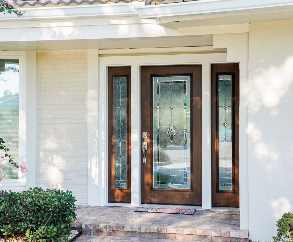 The front entrance boasts an Entry Door St. Louis would be proud of, featuring a dark wooden door with decorative glass panels and sidelights. The house exterior is painted white, with a light brick walkway leading to the inviting entryway, surrounded by lush greenery.