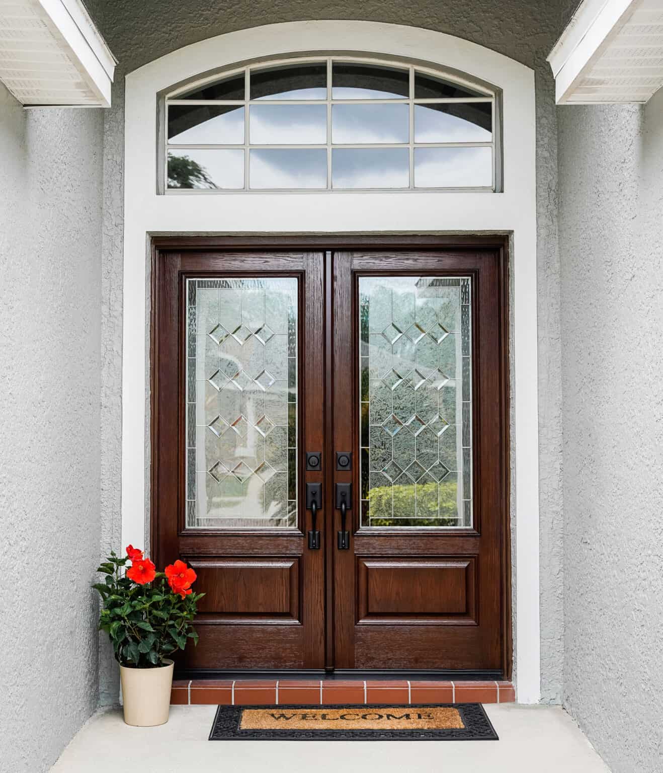 A pair of wooden double doors with decorative glass panels sets a warm tone for Entry Doors St. Louis. A "Welcome" mat graces the brick step, while a potted plant with red flowers adds charm to the left. An arched window elegantly crowns these inviting exterior doors.