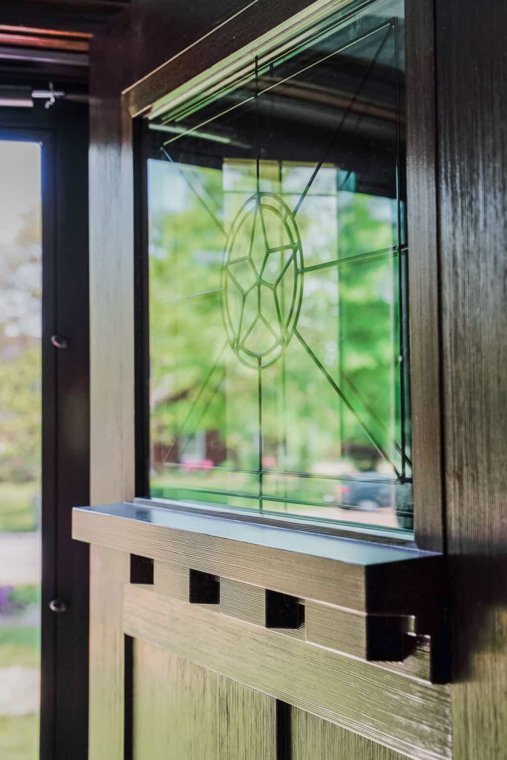 Close-up of an entry door in St. Louis with an intricate stained glass window featuring a geometric pattern. Sunlight illuminates the glass, casting soft reflections, while lush green trees are visible through this elegant exterior door.