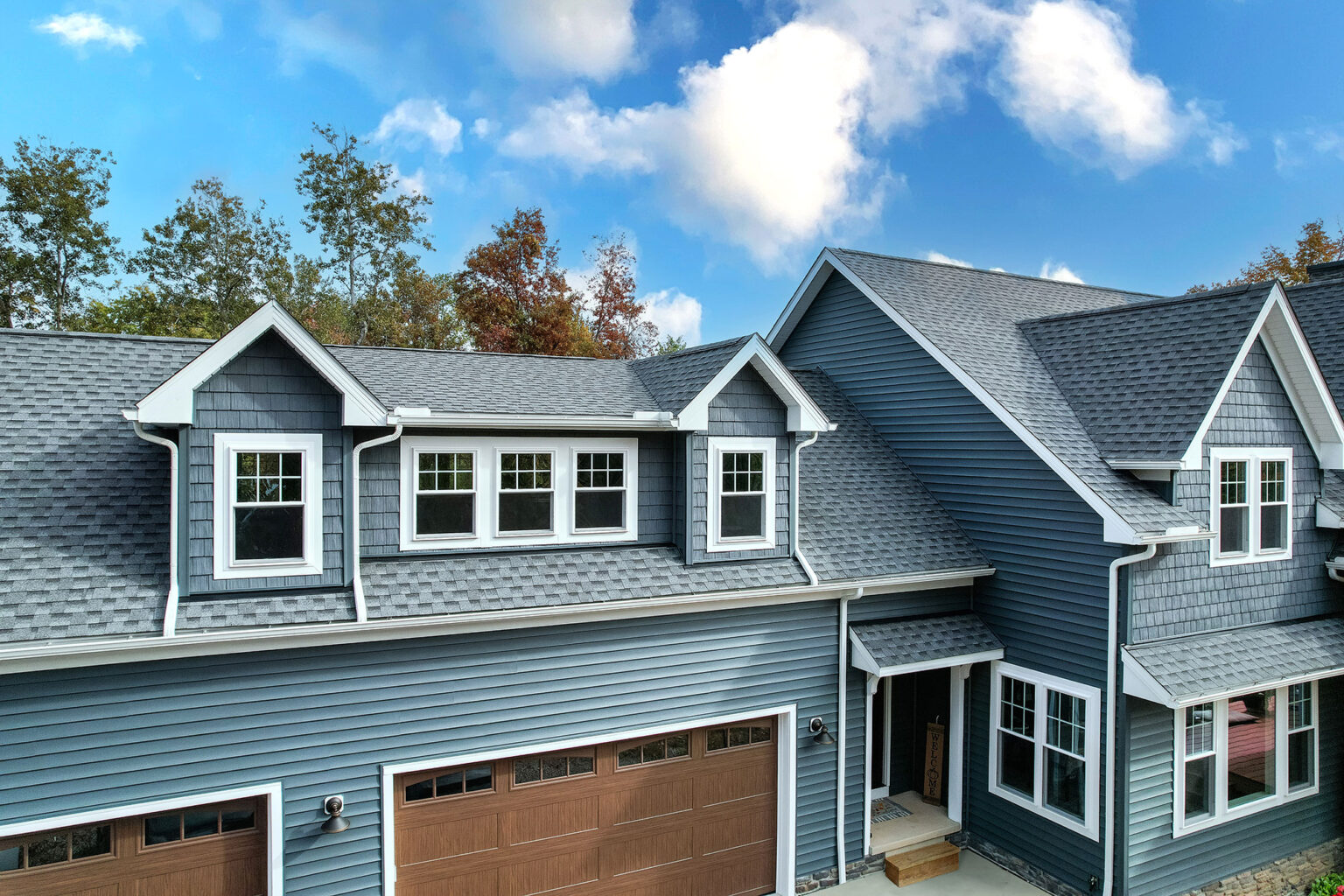 A modern two-story house with blue siding features multiple gabled dormer and double-hung windows. The roof is covered with gray shingles. Brown double garage doors are visible, and trees are in the background under a partly cloudy St. Louis sky.