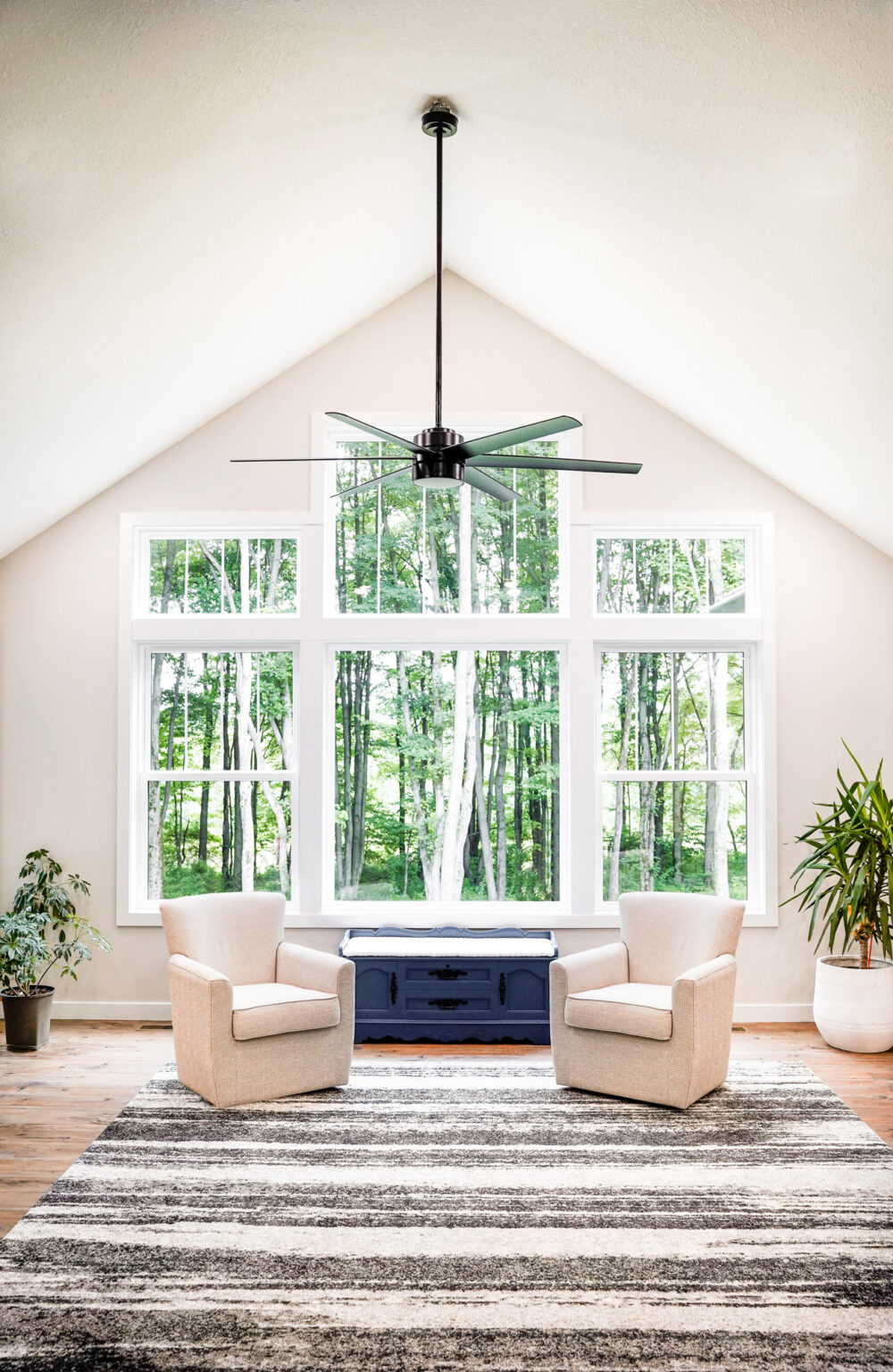 A bright living room with a vaulted ceiling features two beige armchairs facing large double-hung windows overlooking green trees. There's a blue cabinet between the chairs, plants on either side, and a striped gray rug on the wooden floor, creating an inviting St. Louis style ambiance.