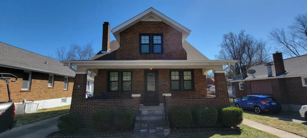 A charming brick house with a steep roof and attic windows. The front features a small porch and a central entrance, with neatly trimmed bushes lining the walkway. It's a sunny day with a clear blue sky.