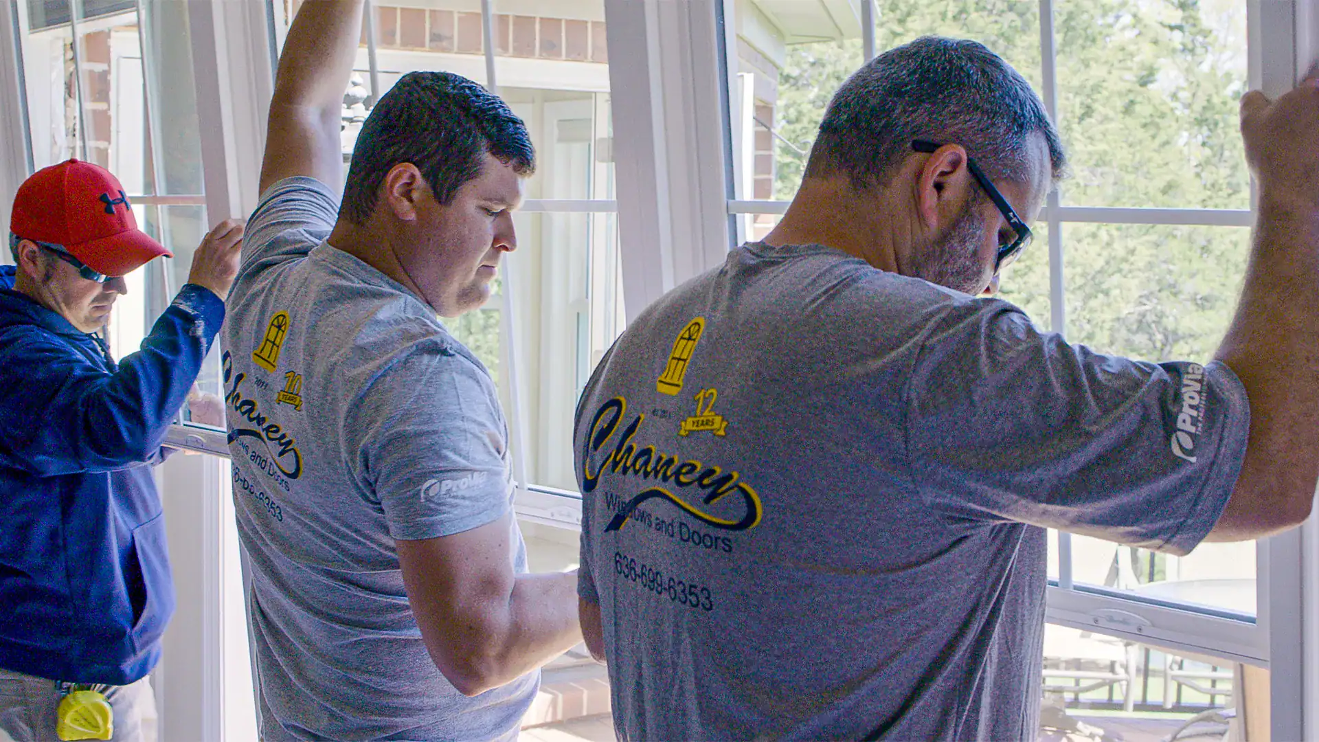 Three men are installing a large window. Two in gray "Chaney" shirts position the window, while a third in a red cap assists on the left. Bright daylight shines through&mdash;hinting that this could be the best time of year to replace your windows & doors.